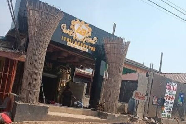 Open-front rustic cafe/restaurant with straw-wrapped pillars and a crest sign above the entrance; sandy ground and power lines overhead.