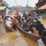 People navigate a flooded street with several wooden boats, loading and unloading goods as neighbors watch; a man wearing an orange life jacket helps transfer items from a boat amid colorful houses on stilts.
