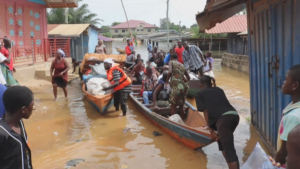 People navigate a flooded street with several wooden boats, loading and unloading goods as neighbors watch; a man wearing an orange life jacket helps transfer items from a boat amid colorful houses on stilts.
