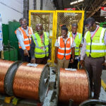 Group of workers in high-visibility vests observing large copper cable spools on the factory floor.
