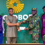 Four men stand in front of a green backdrop as a civilian hands a cheque to a uniformed officer while two other men look on, during a formal presentation event.