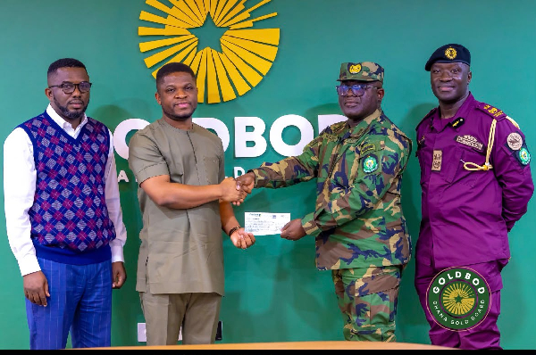 Four men stand in front of a green backdrop as a civilian hands a cheque to a uniformed officer while two other men look on, during a formal presentation event.
