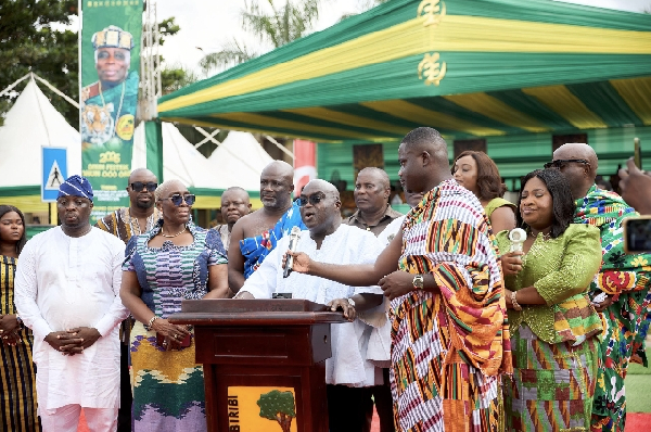 Group of people at an outdoor ceremony around a wooden podium, a man in white speaking to the crowd under a green-yellow canopy.