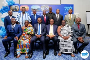 Group portrait of business professionals at a company event, seated and standing in suits and traditional attire with a blue backdrop and logo in the corner.