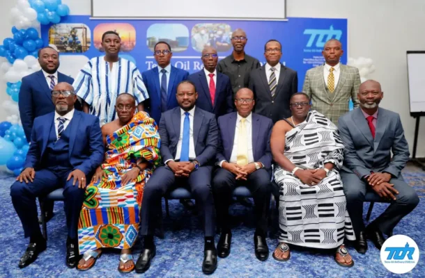 Group portrait of business professionals at a company event, seated and standing in suits and traditional attire with a blue backdrop and logo in the corner.