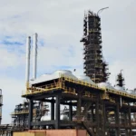 Industrial refinery with tall processing tower, scaffolding, and network of pipes against a cloudy sky, highlighting industrial infrastructure.