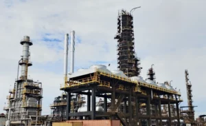 Industrial refinery with tall processing tower, scaffolding, and network of pipes against a cloudy sky, highlighting industrial infrastructure.