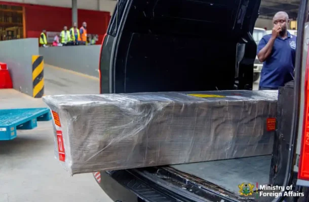75a7c39f-32bf-467d-8e95-7e82820c4e18 | Ghanaguardian.com Large, plastic-wrapped pallet being loaded into the back of an SUV at a loading dock, with ministry logo visible in the corner.