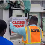 Three workers in high-visibility vests place a 'CLOSED DOWN' sign on a large metal utility cabinet outside a building, signaling a shutdown.