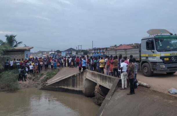 81d86755-f2ac-428a-9b98-4621c822bf05 | Ghanaguardian.com Crowd gathers on a dusty road beside a damaged concrete bridge over a muddy canal, with a large truck on the right labeled EDEKA/Mercedes.