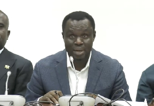 88077481295 | Ghanaguardian.com Man in a blue blazer speaks into a microphone at a panel, seated at a table with others nearby.