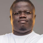 Close-up portrait of a man with a neutral expression wearing a white t-shirt against a gray background.