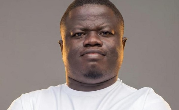 Close-up portrait of a man with a neutral expression wearing a white t-shirt against a gray background.