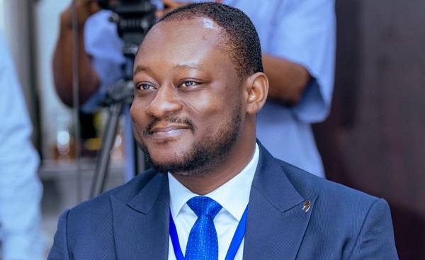96925474 | Ghanaguardian.com Man in a dark blue suit and bright blue tie with a lanyard, seated at an event with a camera in the background.