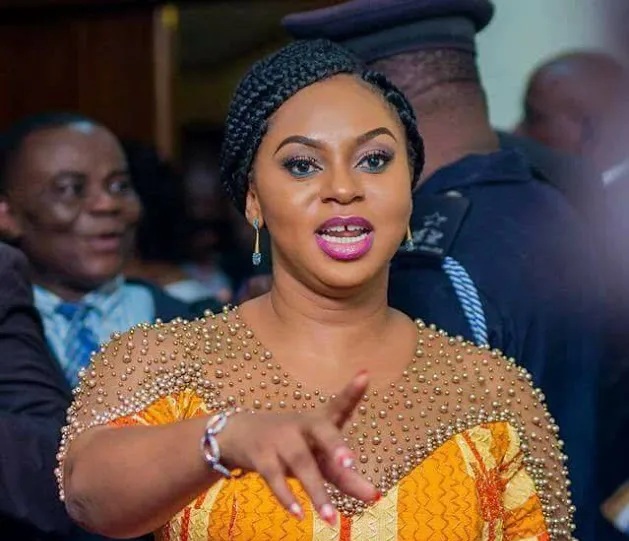 Woman wearing a beaded gold dress and braided headwrap speaks and gestures toward the audience at a crowded event.