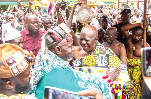 Two men in vibrant traditional attire greet each other at an outdoor ceremony, surrounded by a cheering crowd and photographers.