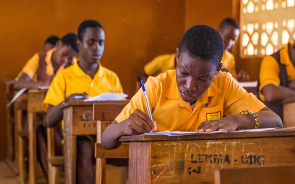 Student in a yellow uniform writes at a wooden desk in a classroom, with peers studying in the background.