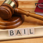 Gavel on a wooden desk with a red binder and glasses; wooden tiles spell 'BAIL' in the foreground.