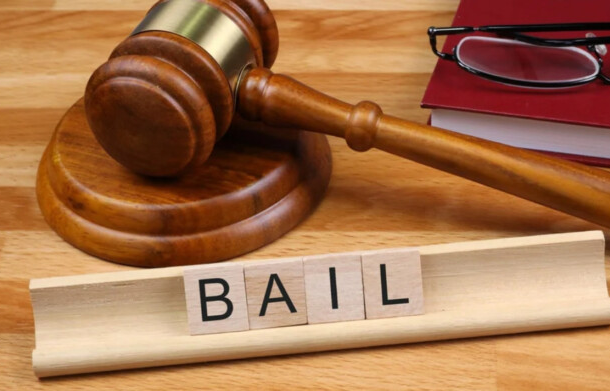 Gavel on a wooden desk with a red binder and glasses; wooden tiles spell 'BAIL' in the foreground.