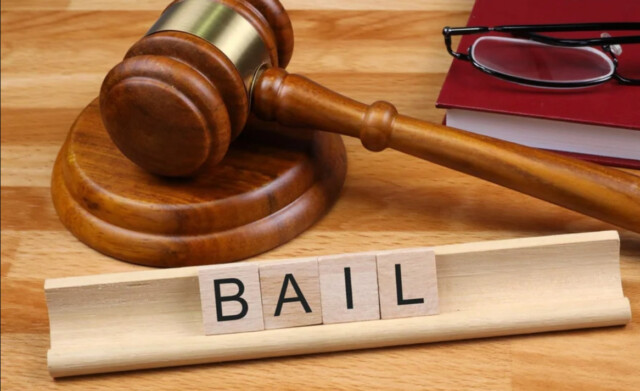 Gavel on a wooden desk with a red binder and glasses; wooden tiles spell 'BAIL' in the foreground.