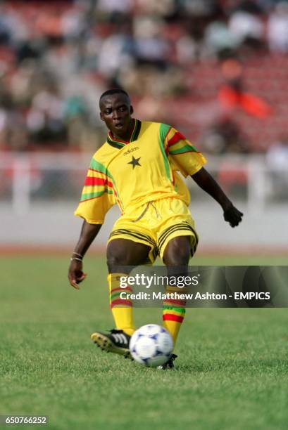 Soccer player in yellow kit dribbling a ball on a grassy field with blurred stands in the background.