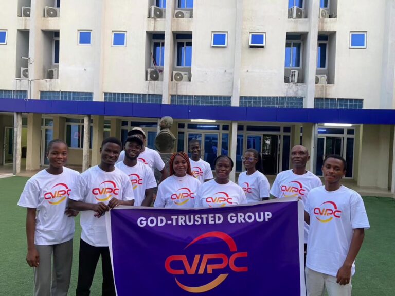 Group of people in white CVPC T-shirts pose outdoors behind a purple banner that reads 'GOD-TRUSTED GROUP' in front of a beige building with blue accents.