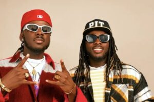 Two men wearing sunglasses pose for a stylish photo: left in a red cap and red jacket making hand signs, right in a PIT cap and striped shirt smiling.