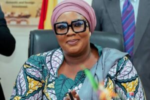 Smiling woman in a pink head wrap and large glasses, wearing a colorful patterned outfit, seated at a desk in a formal meeting setting with flags in the background.
