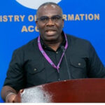 Man speaks at a wooden podium during a Ministry of Information event, wearing a black shirt and a purple lanyard, with a blue backdrop behind him.