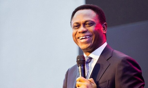 Smiling man in a dark suit speaking into a handheld microphone on stage