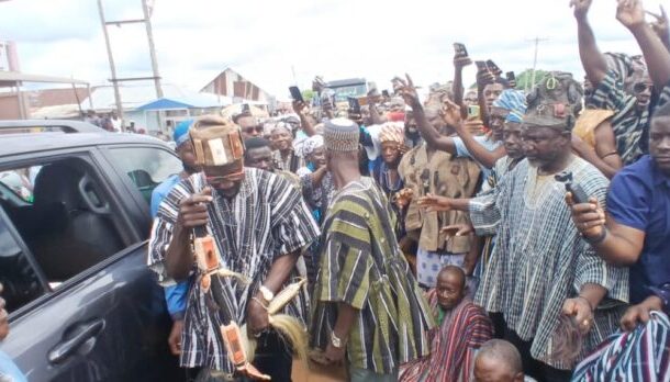 Crowd of people surrounding a man in traditional striped clothing stepping from a car; many hold up phones to take photos and videos.