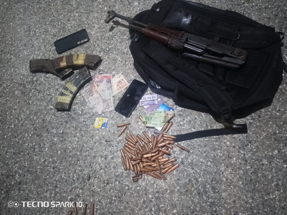 Assault rifle on a black backpack with scattered cash, ammunition, a phone, and cards on a rough floor surface; items appear seized or prepared for transport.