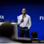 Man in a checkered shirt speaks into a handheld microphone on a stage with FIFA logos in the background.