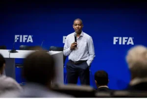Man in a checkered shirt speaks into a handheld microphone on a stage with FIFA logos in the background.