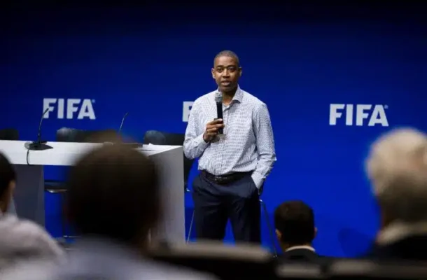 IMG-20260630-WA0018-768x517jpg | Ghanaguardian.com Man in a checkered shirt speaks into a handheld microphone on a stage with FIFA logos in the background.