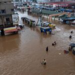 Flooded street market with brown muddy water, vendors and shoppers wading among submerged stalls and awnings.