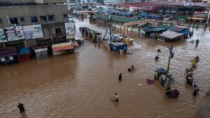Flooded street market with brown muddy water, vendors and shoppers wading among submerged stalls and awnings.