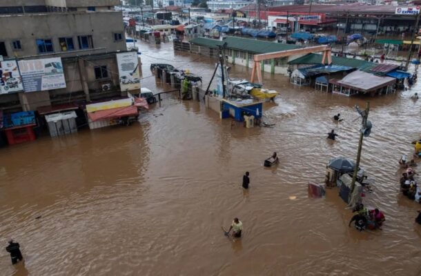 Flooded street market with brown muddy water, vendors and shoppers wading among submerged stalls and awnings.