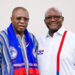 Two men posing for a photo, one in a blue scarf with party logos and the other in a white shirt with red suspenders and a campaign badge.