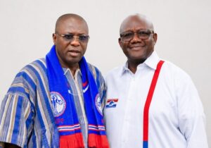 Two men posing for a photo, one in a blue scarf with party logos and the other in a white shirt with red suspenders and a campaign badge.