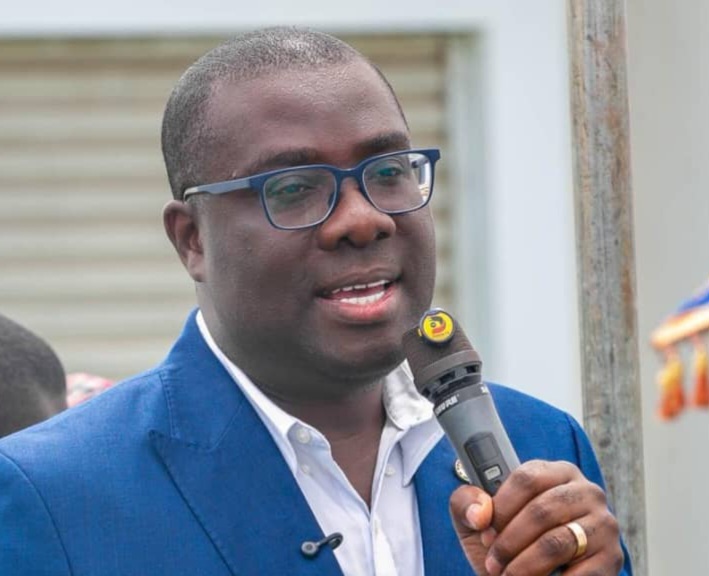 Man in a blue suit and white shirt speaks into a handheld microphone at an outdoor event (public speaking).