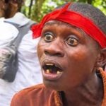 Close-up of a man wearing a bright red headband, with a shocked expression and missing teeth, outdoors in a wooded area.