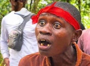 Close-up of a man wearing a bright red headband, with a shocked expression and missing teeth, outdoors in a wooded area.