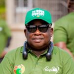 Man in a green polo shirt and sunglasses at an outdoor event, wearing a headset around his neck. The cap reads '24 HOUR ECONOMY' and there are other people in green shirts in the background.