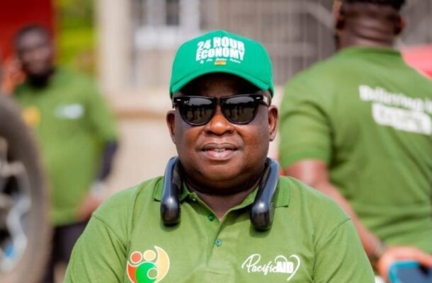 IMG-20260706-WA0000 | Ghanaguardian.com Man in a green polo shirt and sunglasses at an outdoor event, wearing a headset around his neck. The cap reads '24 HOUR ECONOMY' and there are other people in green shirts in the background.