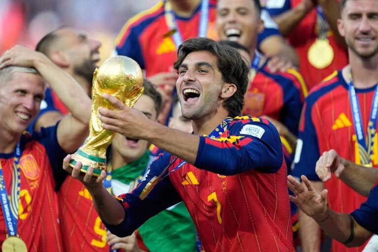 Soccer player in a red Spain jersey lifts the World Cup trophy as teammates celebrate around him.