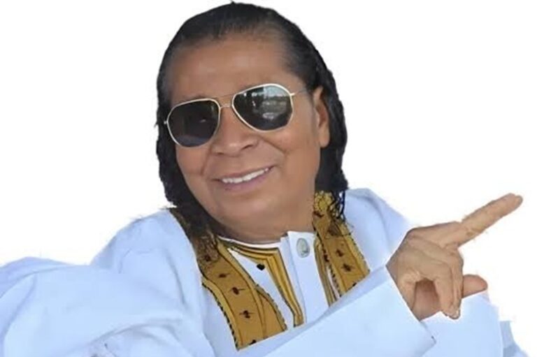 Smiling person wearing sunglasses, a white outfit with a yellow patterned scarf, pointing to the right.