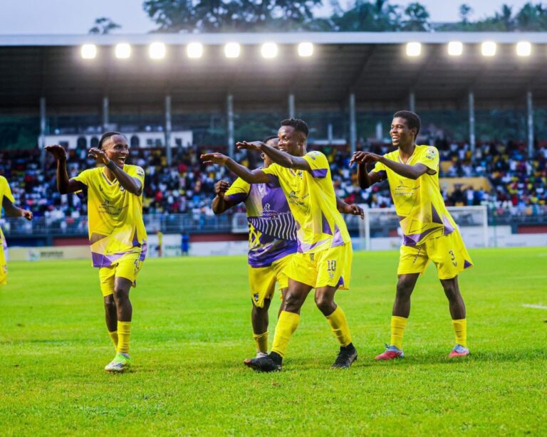 Soccer players in yellow uniforms celebrate on the green field as fans cheer in the stands behind them.