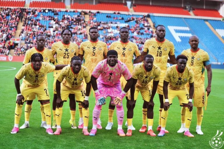 Soccer team in yellow kits posing for a photo on the field with a pink-clad goalkeeper, stadium stands in the background