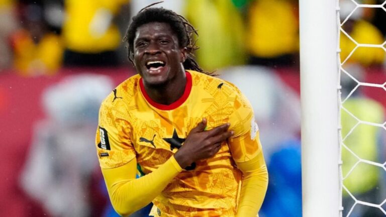 Soccer goalkeeper in a bright yellow jersey celebrates, hand to chest near the goalpost with a cheering crowd behind him.